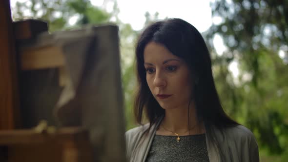 Closeup of a Beautiful Brunette Girl Painting a Landscape on Canvas at an Easel alt