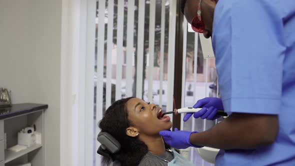 Young Male African Doctor Dentist Fixing and Drying Dental Fillings with a Special Light Holding a alt