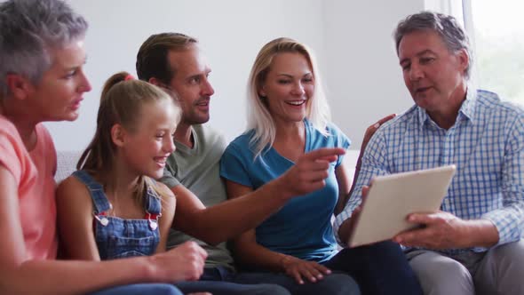 Smiling caucasian parents, grandparents and granddaughter sitting on couch looking at tablet alt