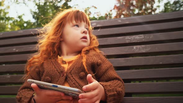 Toddler Girl with Down Syndrome Sitting on the Bench and Playing on Smartphone alt