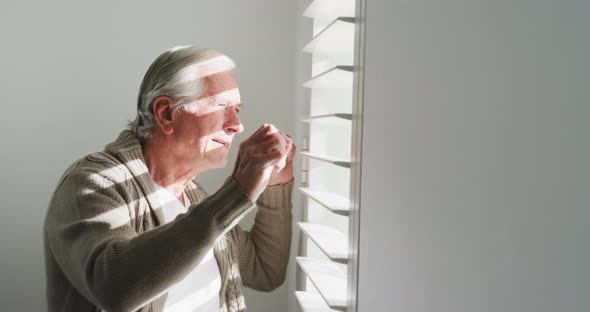 Side view of retired senior man in cardigan sweater looking out through window blinds at home alt