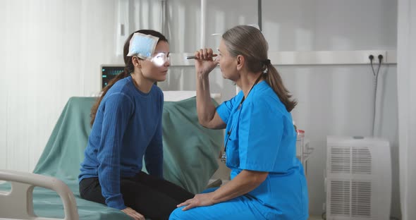 Injured Woman Sitting on Hospital Bed with Doctor Woman Shining in Her Eyes with Flashlight alt