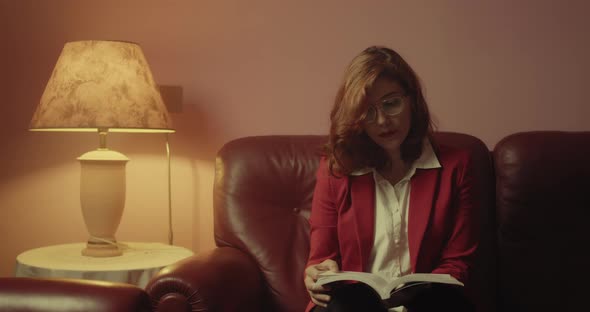 Beautiful woman with red jacket reads the book sitting on the sofa at home with eyeglasses alt