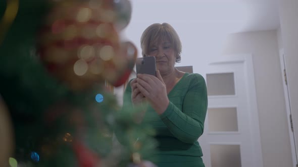 Female Pensioner In The House Capturing Photo Of Glittering Christmas Tree With Cellphone Camera alt