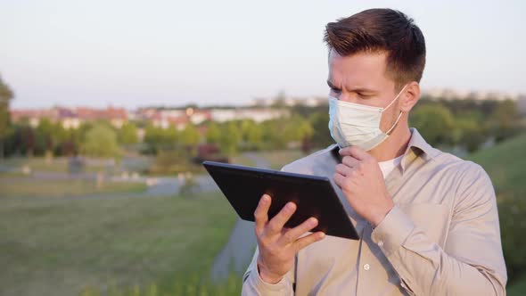 A Young Caucasian Man in a Face Mask Works on a Tablet on a Town's Outskirts alt