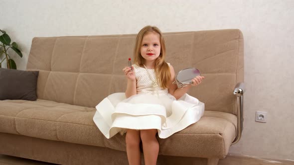A Young Blonde Little Girl Holds a Mirror and Lipstick in Her Hands alt