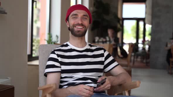 Nice guy with a beard is sitting in a cozy cafe. The man looks at the camera, smiles sincerely alt