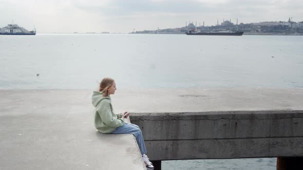 Child with Smartphone Sitting on Sea Pier and Swaying Legs alt
