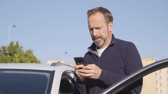 A Middleaged Handsome Caucasian Man Works on a Smartphone As He Stands Next to His Car