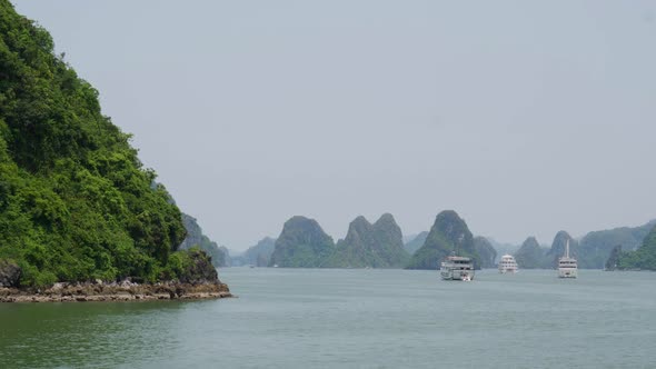 Forested cliff and cruising boats with limestone karsts in the background in HaLong Bay Vietnam alt
