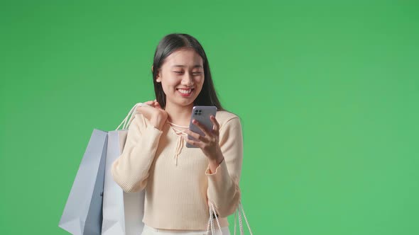 Asian Shopping Woman Holding Shopping Bags Use Mobile Phone And Smile While Walking On Green Screen alt