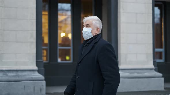 Graying Old Man in Black Coat Walks Near Building in Covid Facemask in Winter