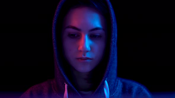 Young Woman in a Hood with a Laptop. Hacker Makes a Hack Through a Laptop. Blue and Red Light Falls alt