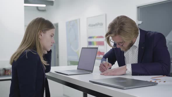 Young Bearded Man in Glasses Wrting Information on the Paper While His Female Colleague Looking at alt