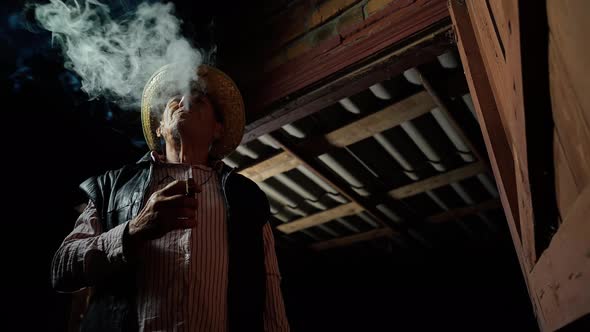 An elderly farmer in a straw hat smokes a cigar outside the ranch. The man smokes a cigar alt