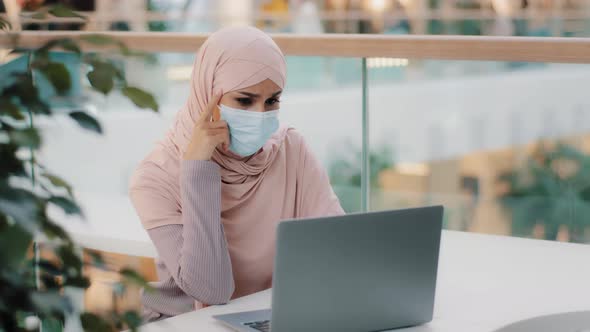 Pensive Young Muslim Woman in Medical Mask in Public Place Sitting Typing on Laptop Arab Girl in alt