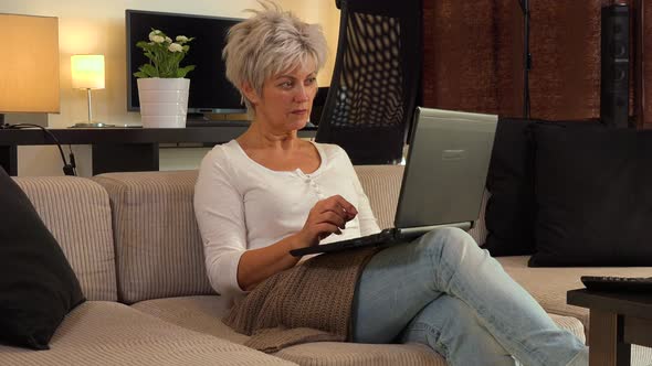A Middle-aged Woman Sits on a Couch in an Apartment Living Room and Works on a Notebook alt