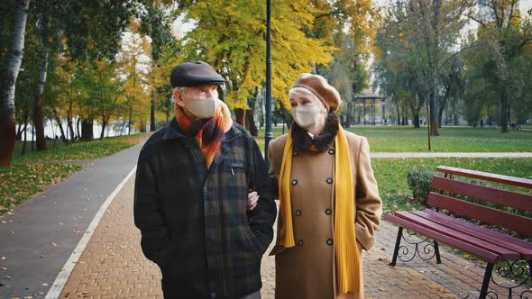 Happy Retired Husband and Wife in Protective Masks are Talking Walking in City Park Near a River alt