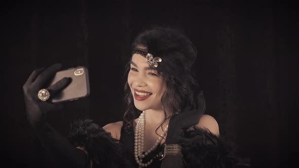 Woman Dressed in Gatsby Style with Boa Making Selfie with Smartphone on Dark alt