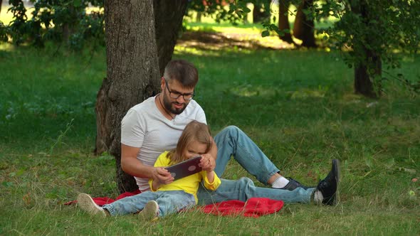 Father and Girl Sit on Lawn in Summer Park with Tablet alt