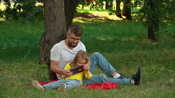 Father and Girl Sit on Lawn in Summer Park with Tablet