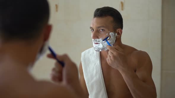 Male With Foam on Face Using Manual Razor to Trim and Shave Beard in Bathroom alt