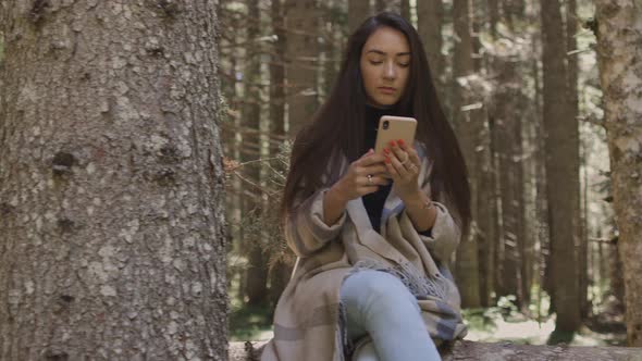 Pretty Woman Using Phone in Forest Sitting on Wooden Log alt