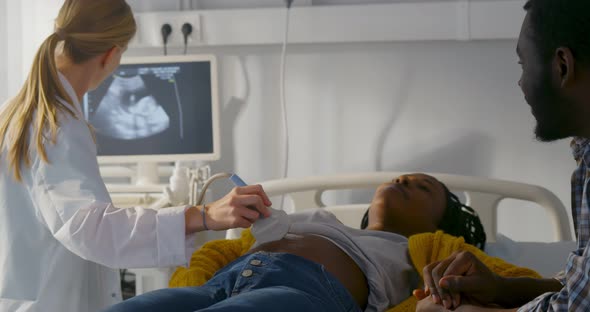 Sonographer Scanning and Examining African Pregnant Woman While Expecting Father Looking at Monitor alt