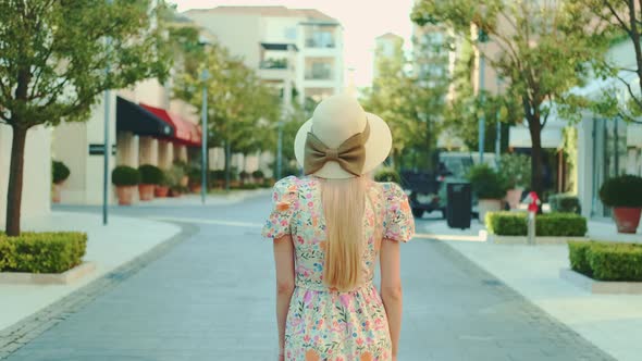 Back View of Slim Blonde Woman in Floral Dress and Summer Hat Walking