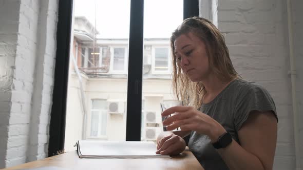 Beautiful Blonde in Gray T-shirt Sits at Table and Reads Magazine. Drinks Water From Transparent alt