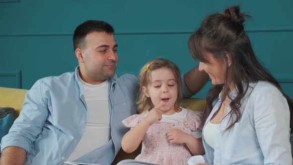 Young Family Reads a Book to Their Little Daughter While Sitting on the Couch alt