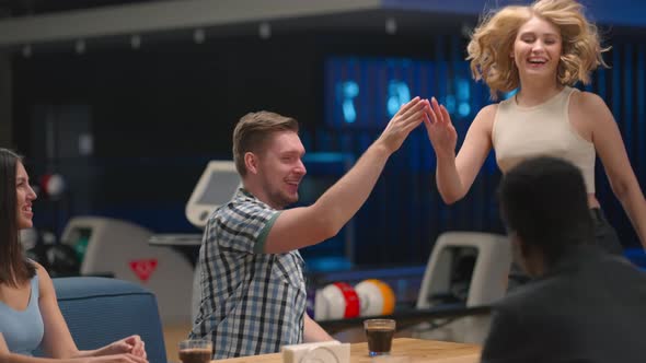 A Multiethnic Group of Friends Sits at a Table Together in a Bowling Alley and Talks Give Five alt