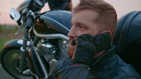 A Middleaged Male Biker Sits By a Motorcycle and Talks on His Mobile Phone