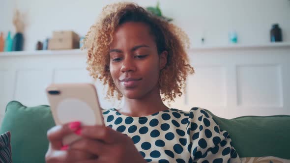 Young Pretty African American Woman with Mobile Phone in Hands Sits on Sofa alt