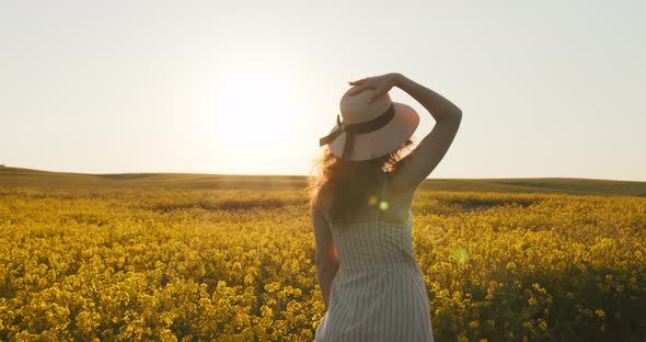 A Beautiful Girl Is Walking Along the Field and Putting on Her Hat alt