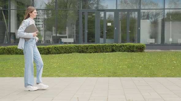 A Girl in a Business Suit with a Laptop in Her Hands Walks Down the Street alt