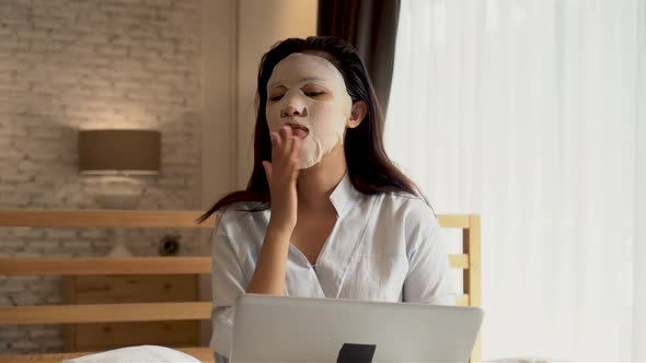 Portrait of Asian Girl Applying Facial Mask on Her Face While Busy Using Computer Laptop in Bedroom alt