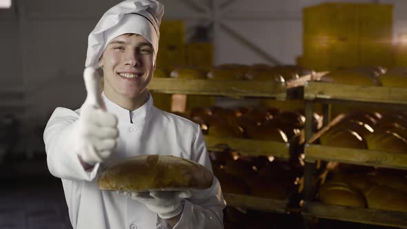 Young Baker Holds a Fresh Loaf of Bread in a Hand and Shows a Thumb at Camera alt
