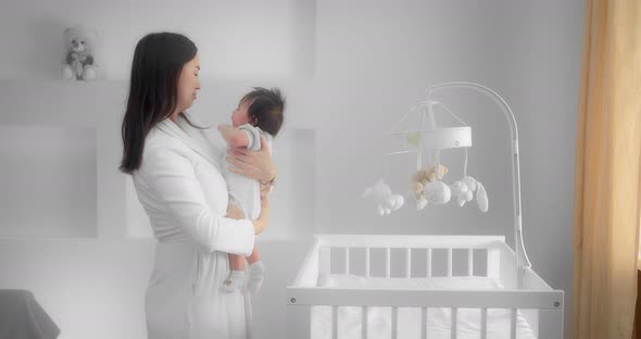 Mother Holds a Newborn Baby in Her Arms and Puts Him in a Child Crib alt