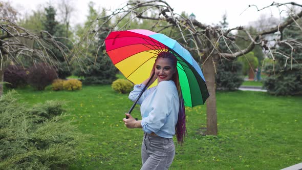 A Cute Girl with Multicolored Braids and Bright Makeup in a Bluish Shirt Posing with a Rainbow alt