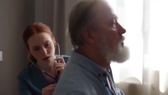 Female Cardiologist Doctor Examining Lungs of Elderly Senior Male Patient at Home Using Stethoscope alt