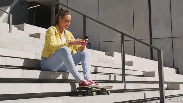 Caucasian woman sitting on stairs with skateboard, using smartphone on sunny day alt