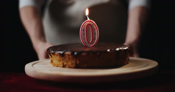 Young Woman Wearing Kitchen Apron Puts Homemade Chocolate Cake or Pie with Number 0 Burning Candle alt