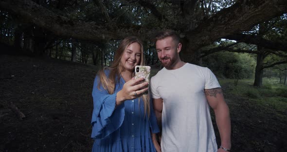 Man and Woman Standing in the Middle of the Forest Looking at Mobile Photos alt