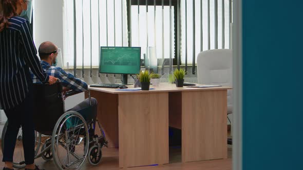 Woman with Protection Mask Helping Disabled Man To Get at Desk alt