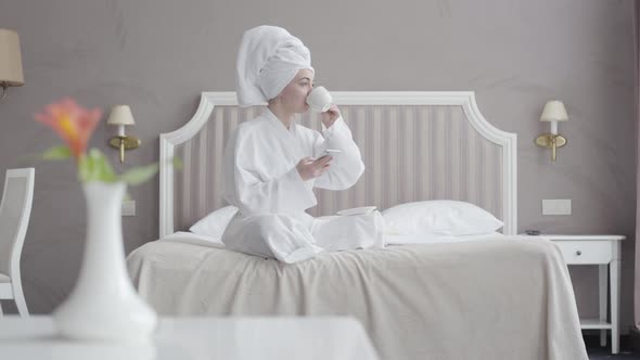 Portrait of Young Caucasian Woman in White Bathrobe and Hair Towel Drinking Coffee and Using Social alt