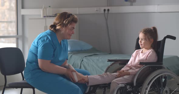 Nurse Massaging Foot of Little Girl Patient Sitting in Wheelchair alt