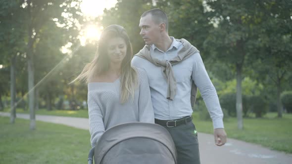 Happy Young Parents Walking with Baby Stroller at Sunset in Summer Park. Portrait of Positive alt
