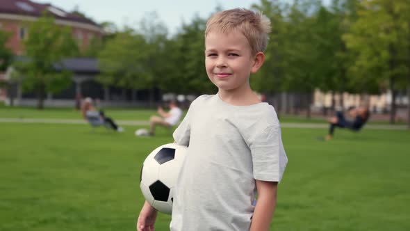 Happily Blonde Preschool Boy Standing in Football Field with Soccer Ball Looking at Camera alt