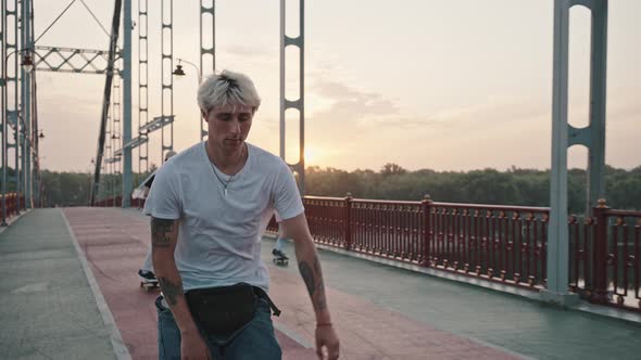 Portrait of a Young Blond Male Skater with Tattoos and a Bright Appearance Riding on the City Bridge alt